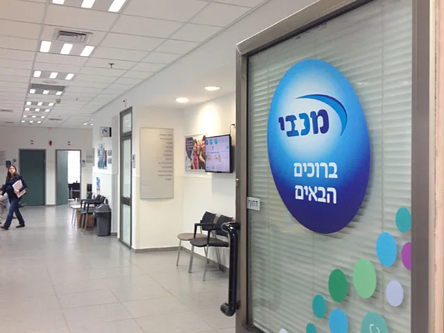 Холл медицинского центра Макаби в Израиле с современным интерьером. На стене логотип клиники Макаби на голубом фоне с надписью на иврите, в коридоре видны стулья для ожидания и проходящие люди